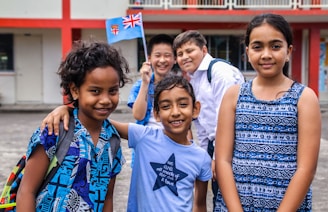 A diverse group of five smiling children standing outdoors near a building with red and white walls. They are wearing colorful clothing, and one child is holding a small flag.