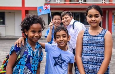 A diverse group of five smiling children standing outdoors near a building with red and white walls. They are wearing colorful clothing, and one child is holding a small flag.