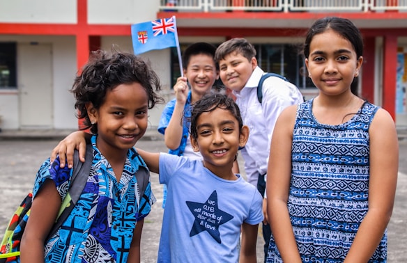 A diverse group of five smiling children standing outdoors near a building with red and white walls. They are wearing colorful clothing, and one child is holding a small flag.