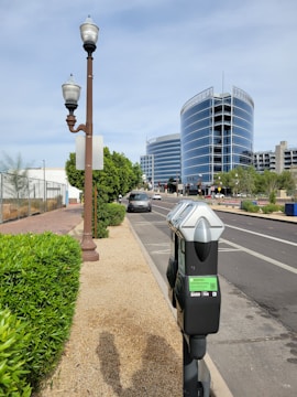 A street scene featuring modern glass buildings with a curved design in the background. In the foreground, there is a parking meter located next to a sidewalk lined with green bushes. The street is relatively empty, with a few cars visible. Street lamps are also present alongside the sidewalk.