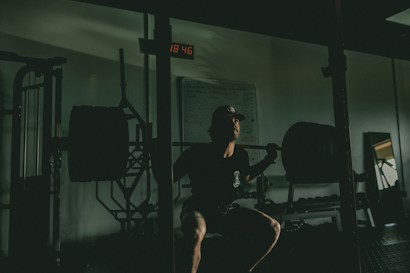 A person is performing a squat in a dimly lit gym environment. They have a heavily loaded barbell across their shoulders. The setting includes gym equipment, such as racks and weights, with a backlit digital clock displaying time on the wall. The ambiance is focused and intense, with shadows accentuating the muscles and form of the individual.