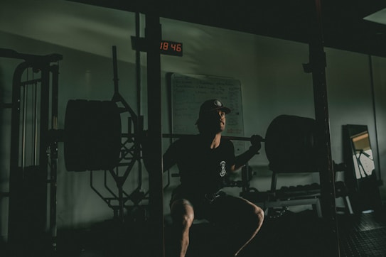 A person is performing a squat in a dimly lit gym environment. They have a heavily loaded barbell across their shoulders. The setting includes gym equipment, such as racks and weights, with a backlit digital clock displaying time on the wall. The ambiance is focused and intense, with shadows accentuating the muscles and form of the individual.