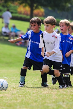 A group of young children are playing soccer on a grassy field. They are wearing soccer uniforms with some in blue and others in white. The children appear to be running toward the ball with expressions of focus and determination. Spectators can be seen blurred in the background.