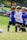 A group of young children are playing soccer on a grassy field. They are wearing soccer uniforms with some in blue and others in white. The children appear to be running toward the ball with expressions of focus and determination. Spectators can be seen blurred in the background.