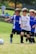 A group of young children are playing soccer on a grassy field. They are wearing soccer uniforms with some in blue and others in white. The children appear to be running toward the ball with expressions of focus and determination. Spectators can be seen blurred in the background.