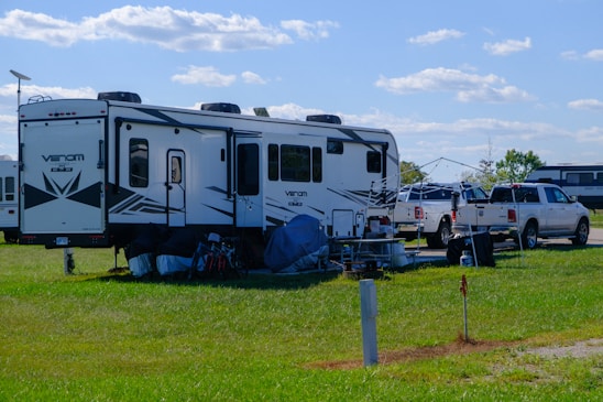 A large white recreational vehicle is parked on a grassy area, accompanied by two white pickup trucks. Various camping gear and chairs are set up near the RV, and the sky is clear with scattered clouds.