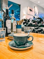 A ceramic coffee cup and saucer with a latte sits on a wooden table, surrounded by glassware, wine bottles, and a utensil holder. The background features potted plants and wall art in a cozy and modern setting.