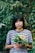 A smiling young woman outdoors surrounded by lush greenery, holding a small potted plant.