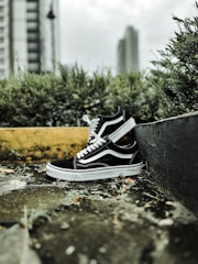 A pair of black and white sneakers is placed on an outdoor stone surface surrounded by green shrubs and a blurred cityscape in the background. The shoes are positioned casually, creating a laid-back and urban vibe.