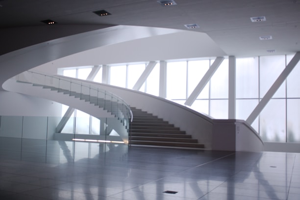 A modern, minimalist interior space featuring a wide, sweeping staircase with glass railings and light streaming in through large, geometrically shaped windows. The floor is made of reflective tiles, enhancing the sense of spaciousness.