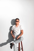 A person with sunglasses and tattoos is sitting on a stool against a plain white background. They are wearing a white t-shirt and light blue jeans, with one leg crossed over the other. The person has a relaxed posture and is holding a cigarette in their hand. The lighting creates a distinct shadow on the wall.