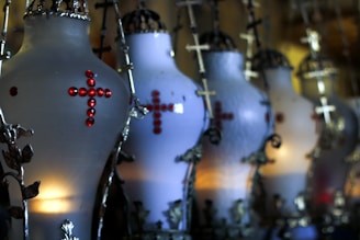 A series of ornate, candle-lit lamps feature intricate metal designs and red gem accents forming a cross shape. The lamps are aligned in a row, projecting a dim, warm light that contrasts with the dark metal elements.