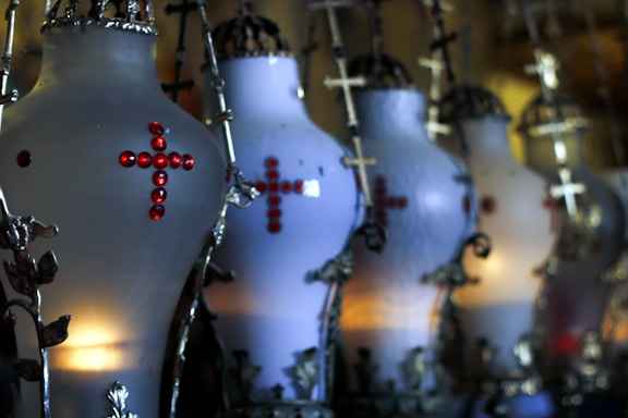 A series of ornate, candle-lit lamps feature intricate metal designs and red gem accents forming a cross shape. The lamps are aligned in a row, projecting a dim, warm light that contrasts with the dark metal elements.