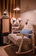 A cozy corner features a vintage reel-to-reel tape recorder placed next to a stack of books and an old-fashioned speaker. A rocking chair with a zigzag patterned fabric and a blue cushion sits underneath a copper-colored floor lamp. The setting exudes a retro and warm ambiance.