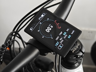 A close-up view of a bicycle handlebar featuring an electronic display with technical readouts. The screen shows information such as speed, distance, and battery percentage. The handlebar and the attached cables are prominently visible, conveying a modern and technical aesthetic.