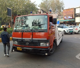 A red and white tow truck is parked on a city street. A man is standing nearby, wearing casual clothing. Several cars are seen in the background, along with a storefront of a pastry shop.