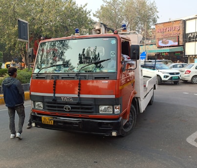 A red and white tow truck is parked on a city street. A man is standing nearby, wearing casual clothing. Several cars are seen in the background, along with a storefront of a pastry shop.
