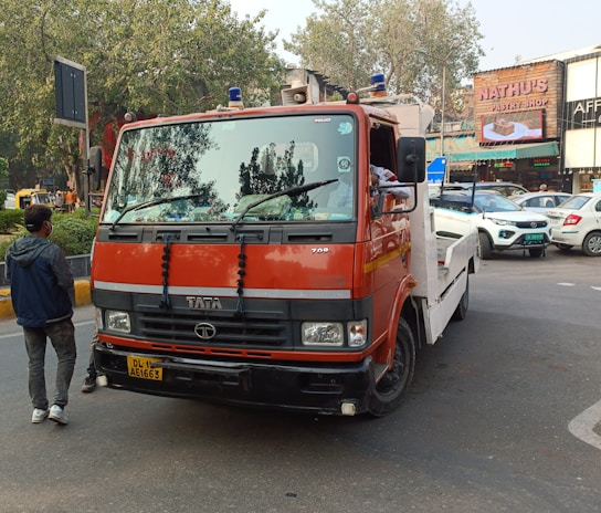A red and white tow truck is parked on a city street. A man is standing nearby, wearing casual clothing. Several cars are seen in the background, along with a storefront of a pastry shop.
