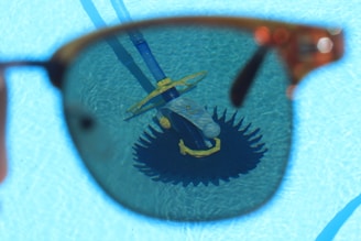 A sunglass lens frames the view of a pool cleaning device submerged in water, creating a contrasting focus against the bright blue of the pool. The lens slightly darkens the view, emphasizing the details of the cleaning equipment with its brush and hose.