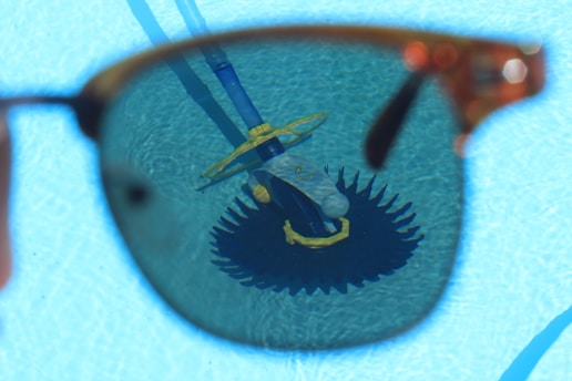 A sunglass lens frames the view of a pool cleaning device submerged in water, creating a contrasting focus against the bright blue of the pool. The lens slightly darkens the view, emphasizing the details of the cleaning equipment with its brush and hose.