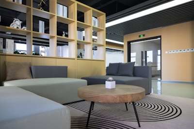 A modern office space featuring a stylish seating area with two sofas in shades of grey and beige. In the foreground, a round wooden coffee table with a small potted plant sits on a patterned rug. Behind the seating area is a wooden shelving unit filled with books and decorative items, leading to a doorway with a glass door. The room is well-lit with natural light coming from the large windows.