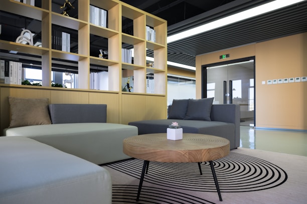 A modern office space featuring a stylish seating area with two sofas in shades of grey and beige. In the foreground, a round wooden coffee table with a small potted plant sits on a patterned rug. Behind the seating area is a wooden shelving unit filled with books and decorative items, leading to a doorway with a glass door. The room is well-lit with natural light coming from the large windows.