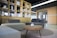 A modern office space featuring a stylish seating area with two sofas in shades of grey and beige. In the foreground, a round wooden coffee table with a small potted plant sits on a patterned rug. Behind the seating area is a wooden shelving unit filled with books and decorative items, leading to a doorway with a glass door. The room is well-lit with natural light coming from the large windows.