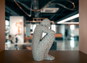 A sculpture composed of small, textured blocks forms a human figure in a seated, contemplative pose. The sculpture is placed in a modern, open indoor space with blurred background elements including abstract art, plants, and contemporary furnishings.
