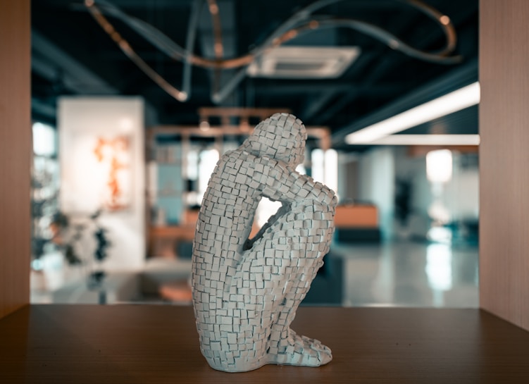 A sculpture composed of small, textured blocks forms a human figure in a seated, contemplative pose. The sculpture is placed in a modern, open indoor space with blurred background elements including abstract art, plants, and contemporary furnishings.
