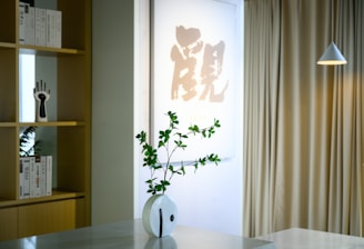 A minimalist interior space featuring a decorative vase with green foliage on a light-colored table. In the background, a shelf displays books and a unique hand sculpture. A piece of Asian calligraphy art is prominently displayed, alongside beige curtains and a modern pendant lamp hanging from the ceiling.