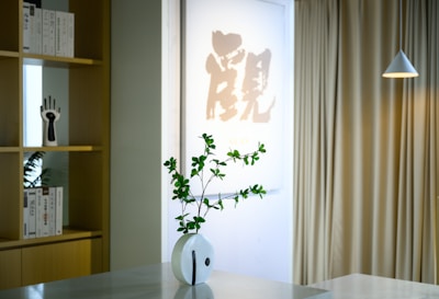 A minimalist interior space featuring a decorative vase with green foliage on a light-colored table. In the background, a shelf displays books and a unique hand sculpture. A piece of Asian calligraphy art is prominently displayed, alongside beige curtains and a modern pendant lamp hanging from the ceiling.