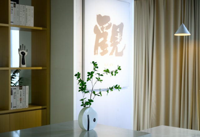 A minimalist interior space featuring a decorative vase with green foliage on a light-colored table. In the background, a shelf displays books and a unique hand sculpture. A piece of Asian calligraphy art is prominently displayed, alongside beige curtains and a modern pendant lamp hanging from the ceiling.