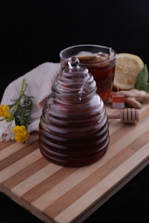 A glass jar with ridged sides filled with dark liquid, likely honey, is placed on a wooden board. Beside it, there is a cup of tea with a lemon slice and a tea bag, accompanied by a piece of ginger and a honey dipper. A white cloth partially covers some flowers, including yellow and white blooms. The background is dark, contrasting with the warm, natural tones of the objects.