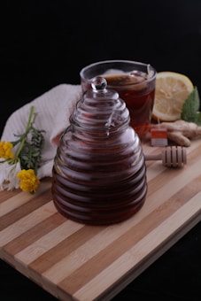 A glass jar with ridged sides filled with dark liquid, likely honey, is placed on a wooden board. Beside it, there is a cup of tea with a lemon slice and a tea bag, accompanied by a piece of ginger and a honey dipper. A white cloth partially covers some flowers, including yellow and white blooms. The background is dark, contrasting with the warm, natural tones of the objects.
