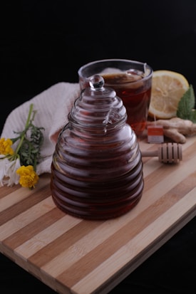 A glass jar with ridged sides filled with dark liquid, likely honey, is placed on a wooden board. Beside it, there is a cup of tea with a lemon slice and a tea bag, accompanied by a piece of ginger and a honey dipper. A white cloth partially covers some flowers, including yellow and white blooms. The background is dark, contrasting with the warm, natural tones of the objects.