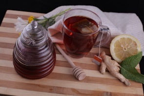 A glass jar filled with honey sits next to a cup of tea with a tea bag submerged inside. A honey dipper rests on the wooden cutting board. Beside the cup, a lemon half, a piece of ginger, and a sprig of mint are arranged. In the background, a light-colored towel and a few flowers are visible.