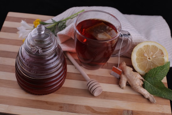 A glass jar filled with honey sits next to a cup of tea with a tea bag submerged inside. A honey dipper rests on the wooden cutting board. Beside the cup, a lemon half, a piece of ginger, and a sprig of mint are arranged. In the background, a light-colored towel and a few flowers are visible.