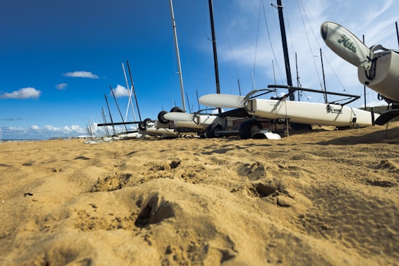 Several catamarans are lined up on a sandy beach under a clear blue sky. The perspective is low to the ground, emphasizing the sandy texture and the rigging and masts of the boats. A few scattered clouds are visible in the distance.