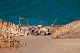 A yellow industrial vehicle is operating at a wood processing facility near a large body of water. Stacks of neatly piled logs are arranged on either side of the vehicle. The water in the background has a vivid blue color.