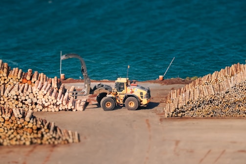 A yellow industrial vehicle is operating at a wood processing facility near a large body of water. Stacks of neatly piled logs are arranged on either side of the vehicle. The water in the background has a vivid blue color.