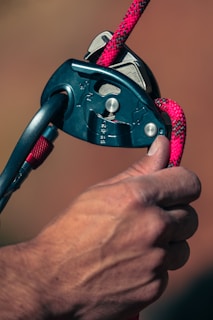 A close-up of a person's hand holding a belay device with a carabiner and a climbing rope. The rope is threaded through the device, which is black with metallic elements, and the rope is red with black patterns.