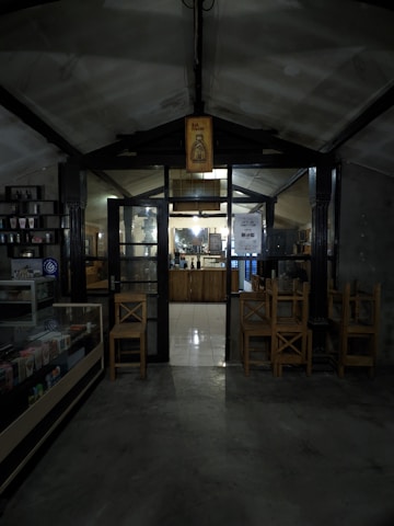 A dimly lit interior of what appears to be a cozy café or small shop. The entrance is framed by dark wooden beams and a glass door, with a sign overhead featuring a bottle illustration and the words 'Hot Promo'. Inside, a counter is visible with shelves and various items displayed. Wooden chairs are stacked nearby, and there is a refrigerated display case on the left.
