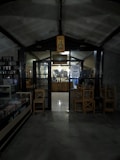 A dimly lit interior of what appears to be a cozy caf&eacute; or small shop. The entrance is framed by dark wooden beams and a glass door, with a sign overhead featuring a bottle illustration and the words 'Hot Promo'. Inside, a counter is visible with shelves and various items displayed. Wooden chairs are stacked nearby, and there is a refrigerated display case on the left.