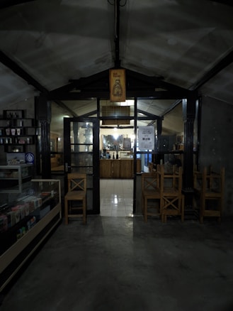 A dimly lit interior of what appears to be a cozy café or small shop. The entrance is framed by dark wooden beams and a glass door, with a sign overhead featuring a bottle illustration and the words 'Hot Promo'. Inside, a counter is visible with shelves and various items displayed. Wooden chairs are stacked nearby, and there is a refrigerated display case on the left.