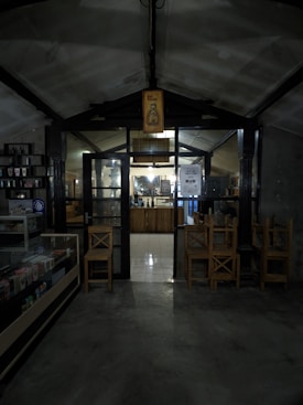 A dimly lit interior of what appears to be a cozy café or small shop. The entrance is framed by dark wooden beams and a glass door, with a sign overhead featuring a bottle illustration and the words 'Hot Promo'. Inside, a counter is visible with shelves and various items displayed. Wooden chairs are stacked nearby, and there is a refrigerated display case on the left.