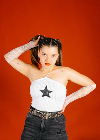A woman stands against a vibrant red background, wearing a white halter top with a black star design. Her hair is styled in pigtails, and she holds one with her hand. She wears black jeans, a belt with circular metal details, and white mesh gloves on her arms. Her expression is confident with bright red lipstick.