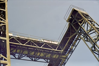 A large, industrial steel structure extends diagonally against a clear sky. The structure appears to be part of an old bridge or crane, with rust and signs of weathering visible on the metal beams.