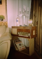 A cozy corner featuring a vintage wooden radio on a simple wooden stand, adorned with a silver candelabra, a globe, and a clear bottle. Beside the radio, there is a collection of vinyl records leaning against the stand. The setting includes a beige leather sofa and soft white curtains, with logs stacked against the wall as part of the decor. A small potted plant adds a touch of greenery to the rustic atmosphere.