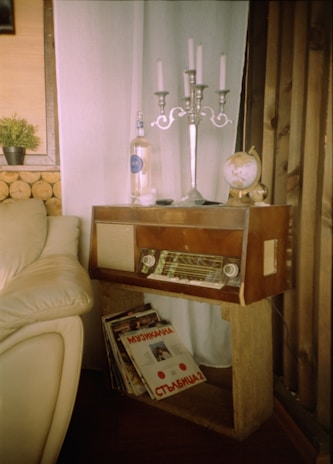 A cozy corner featuring a vintage wooden radio on a simple wooden stand, adorned with a silver candelabra, a globe, and a clear bottle. Beside the radio, there is a collection of vinyl records leaning against the stand. The setting includes a beige leather sofa and soft white curtains, with logs stacked against the wall as part of the decor. A small potted plant adds a touch of greenery to the rustic atmosphere.
