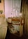 A cozy corner featuring a vintage wooden radio on a simple wooden stand, adorned with a silver candelabra, a globe, and a clear bottle. Beside the radio, there is a collection of vinyl records leaning against the stand. The setting includes a beige leather sofa and soft white curtains, with logs stacked against the wall as part of the decor. A small potted plant adds a touch of greenery to the rustic atmosphere.